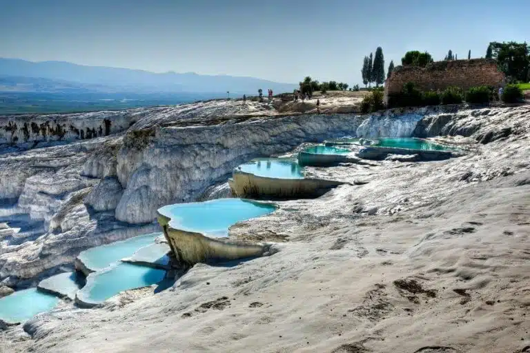 View of the terraced thermal pools of Hierapolis-Pamukkale in Turkey, showcasing bright white limestone formations filled with mineral-rich turquoise waters under a clear blue sky, with ancient Greco-Roman ruins visible in the background.