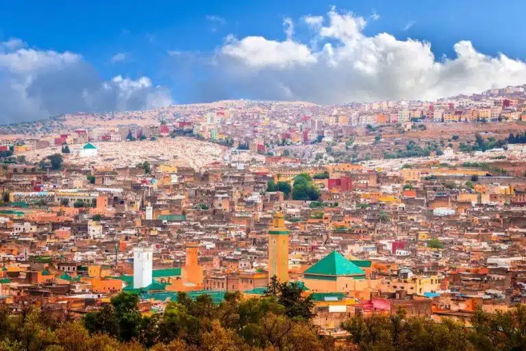 A panoramic view of Marrakech city showing a vibrant landscape of terracotta-colored buildings, mosques, and the distant Atlas Mountains under a clear sky.